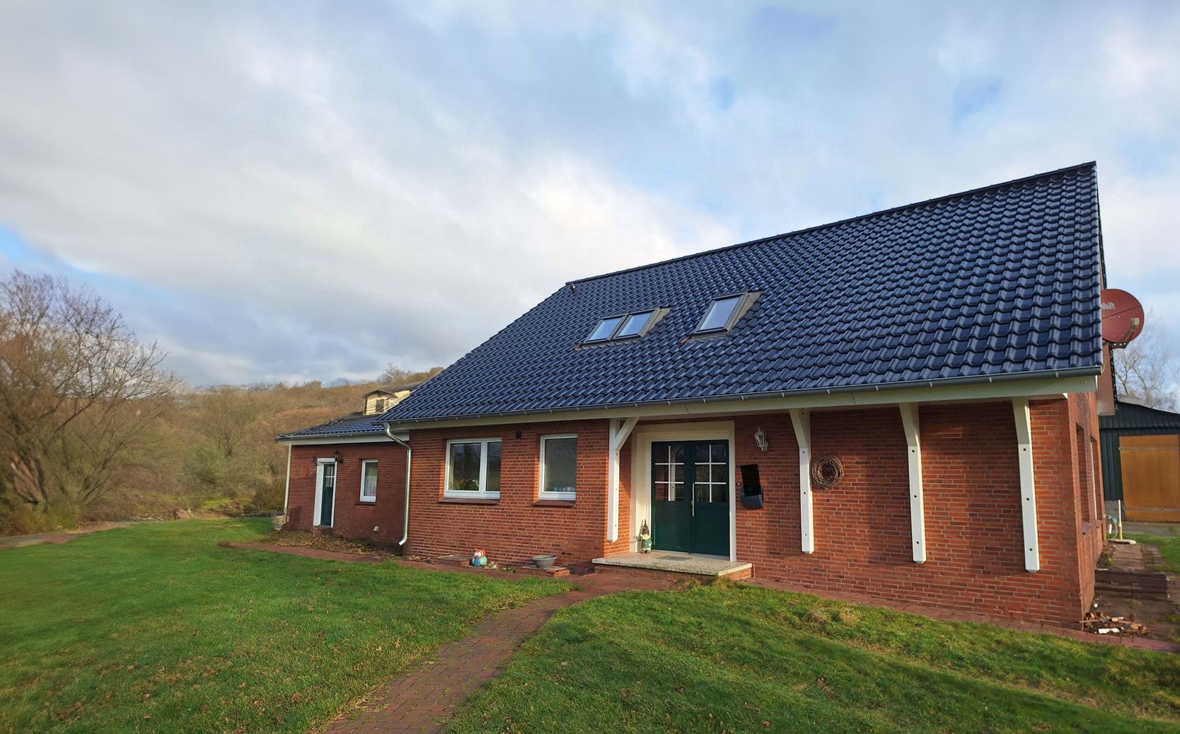 Ein Backsteinhaus mit einem dunkelblauen Ziegeldach, einer grünen Tür, grünen Fenstern und einem Seitengarten vor einem bewölkten Himmel.