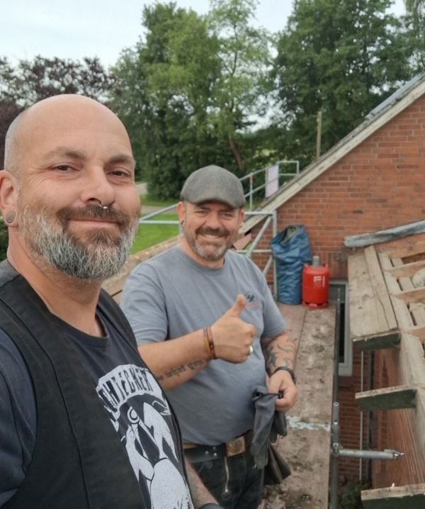 Zwei Männer posieren auf einem Dach, einer zeigt den Daumen nach oben, im Hintergrund sind Grünpflanzen und ein roter Container zu sehen.