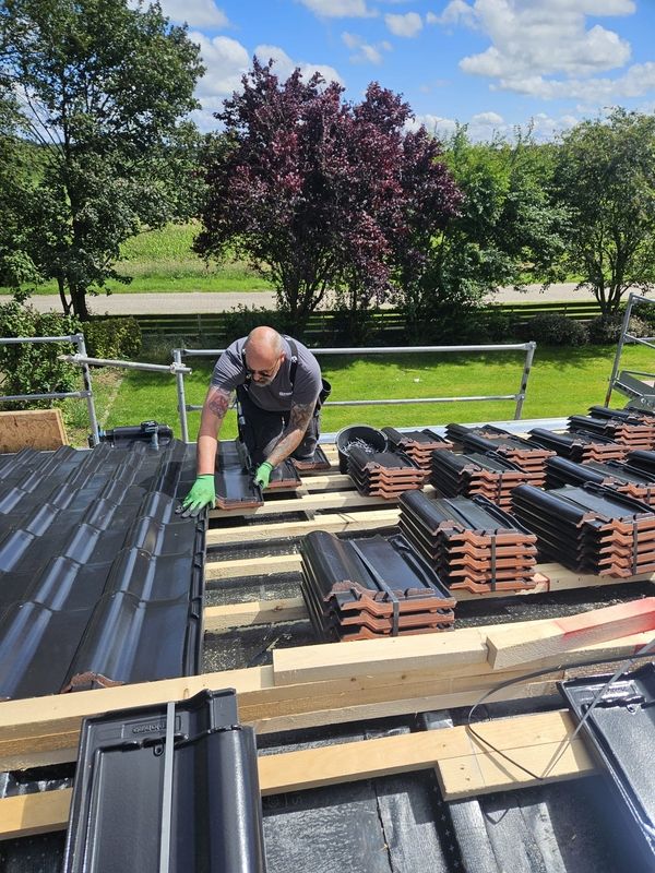 Ein Arbeiter mit grünen Handschuhen installiert schwarze Dachziegel auf einem Holzrahmen, umgeben von gestapelten Ziegeln und grünen Bäumen unter einem blauen Himmel.