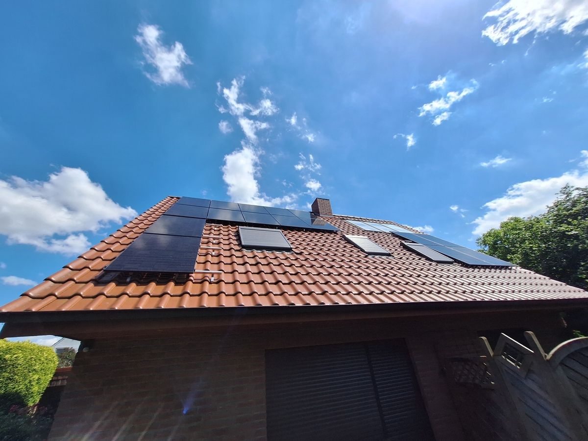 Ein schräges braunes Ziegeldach mit Solarmodulen unter einem strahlend blauen Himmel mit vereinzelten Wolken. In der Nähe sind grüne Bäume zu sehen.