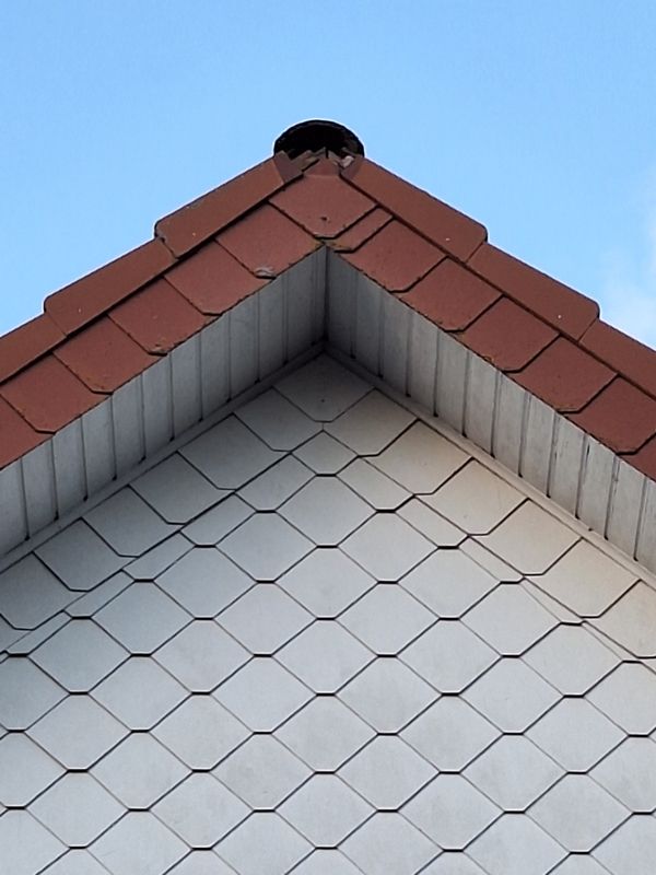 Blick auf den Giebel eines Hausdachs mit roten Schindeln und einem weißen sechseckigen Muster, mit einer runden Öffnung an der Spitze vor einem blauen Himmel.
